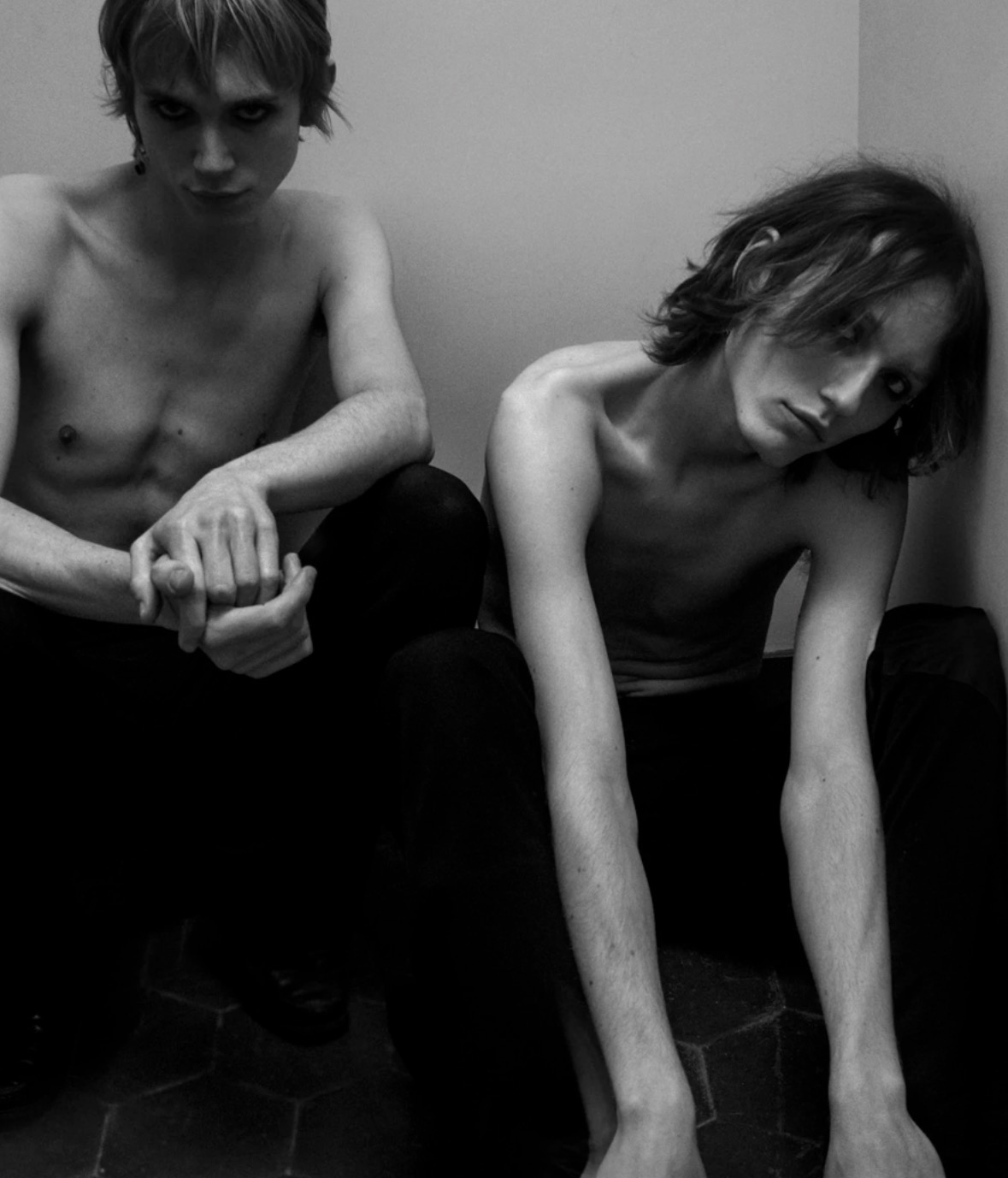 The two members of POL seated against a wall in a black-and-white portrait.